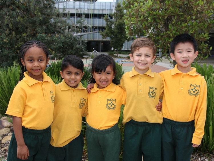 Five young students in yellow school uniforms standing outdoors, smiling with their arms around each other, in front of trees and school buildings.